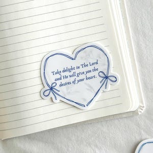 May include: A white heart-shaped sticker with a blue outline and blue ribbon accents. The sticker has a marble-like background and features the text "Take delight in The Lord and He will give you the desires of your heart."