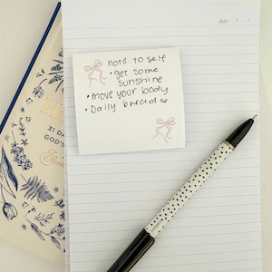 Peut inclure: Un carnet &agrave; spirales dor&eacute;es &agrave; lignes blanches. Un post-it avec le texte "note to self &bull; get some sunshine &bull; move your body &bull; Daily bread" est coll&eacute; sur le carnet. Un stylo noir &agrave; pois blancs est pos&eacute; sur le carnet.