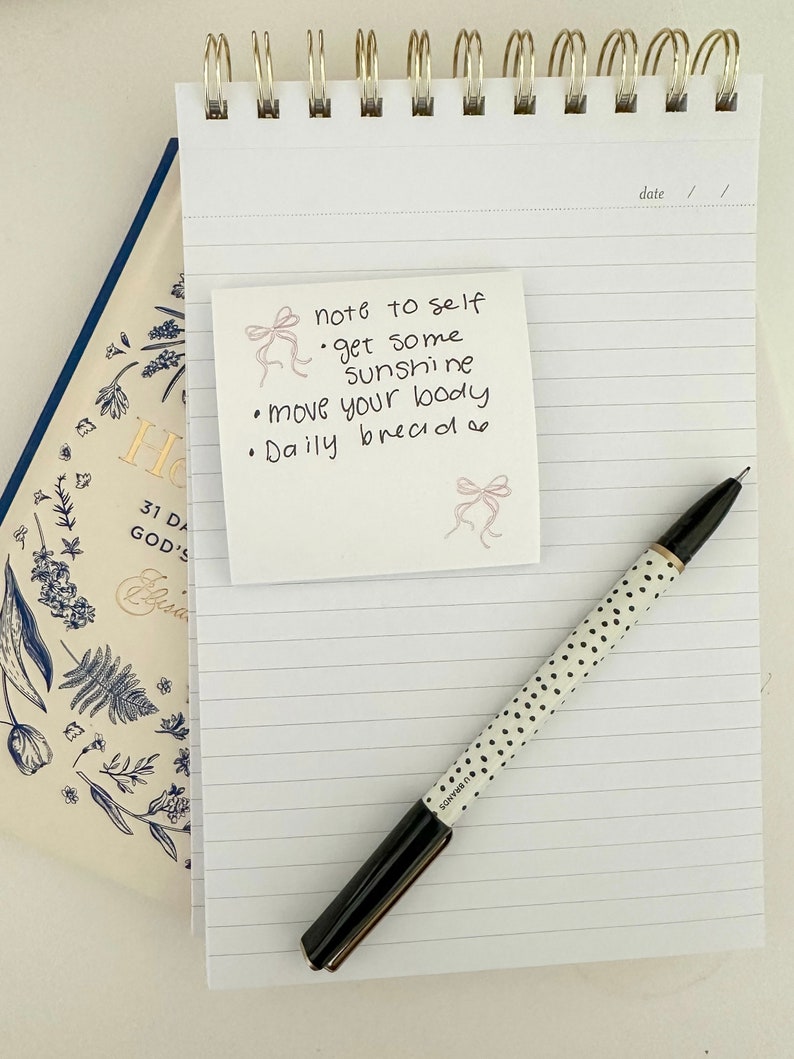 Peut inclure: Un carnet &agrave; spirales dor&eacute;es &agrave; lignes blanches. Un post-it avec le texte "note to self &bull; get some sunshine &bull; move your body &bull; Daily bread" est coll&eacute; sur le carnet. Un stylo noir &agrave; pois blancs est pos&eacute; sur le carnet.