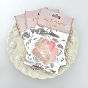 May include: Air fresheners in silver packaging with a floral design and the text "Find your worth in God." The air fresheners are on a white, floral-patterned plate. The scent is "Cotton Candy."
