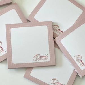 May include: A stack of pink sticky notes with a white square in the centre and the text "Thankful Grateful Blessed" in a pink and red font.