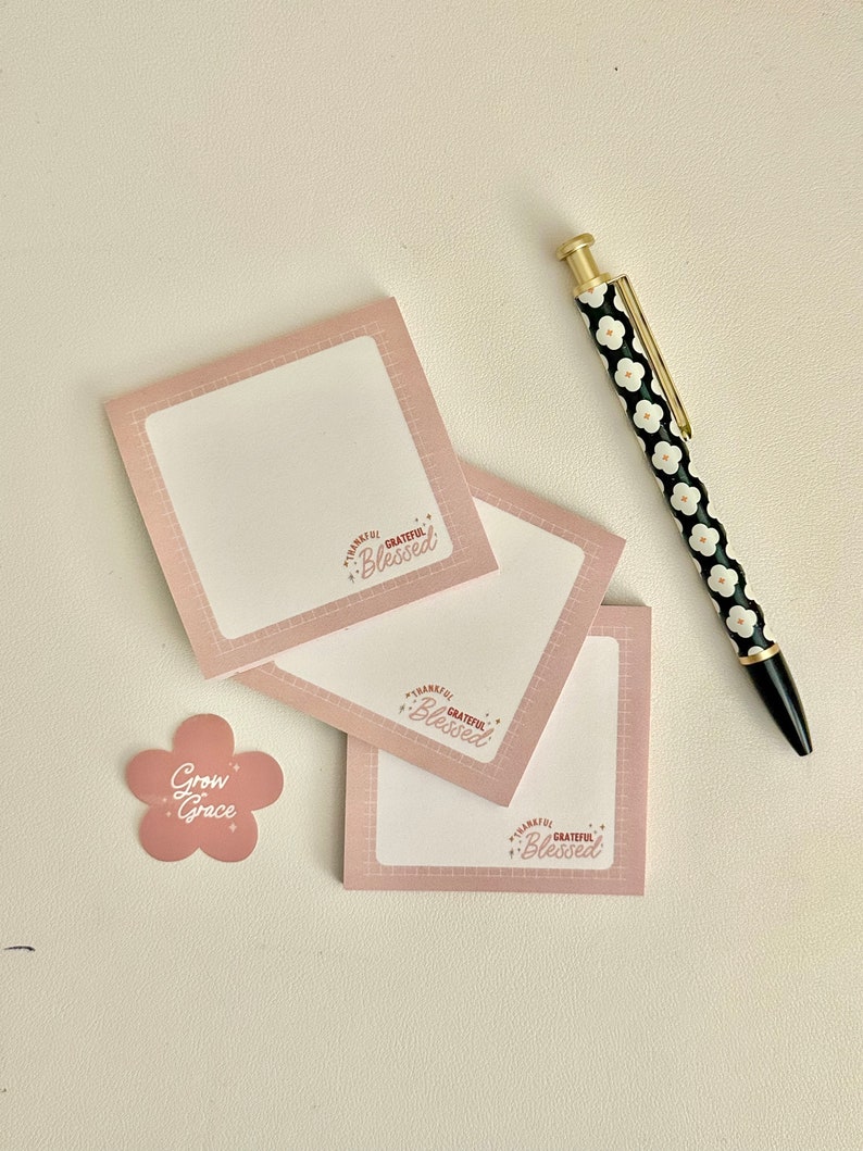May include: Three pink square sticky notes with the text "Thankful Grateful Blessed" in a white circle with stars. A pink flower sticker with the text "Grow Grace" and a black and white pen with a floral pattern.