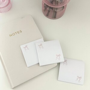 Peut inclure: Trois blocs de notes autocollantes carr&eacute;s blancs avec un motif de n&oelig;ud rose. Les blocs de notes sont sur une surface blanche avec un carnet blanc et un vase en verre rose en arri&egrave;re-plan.