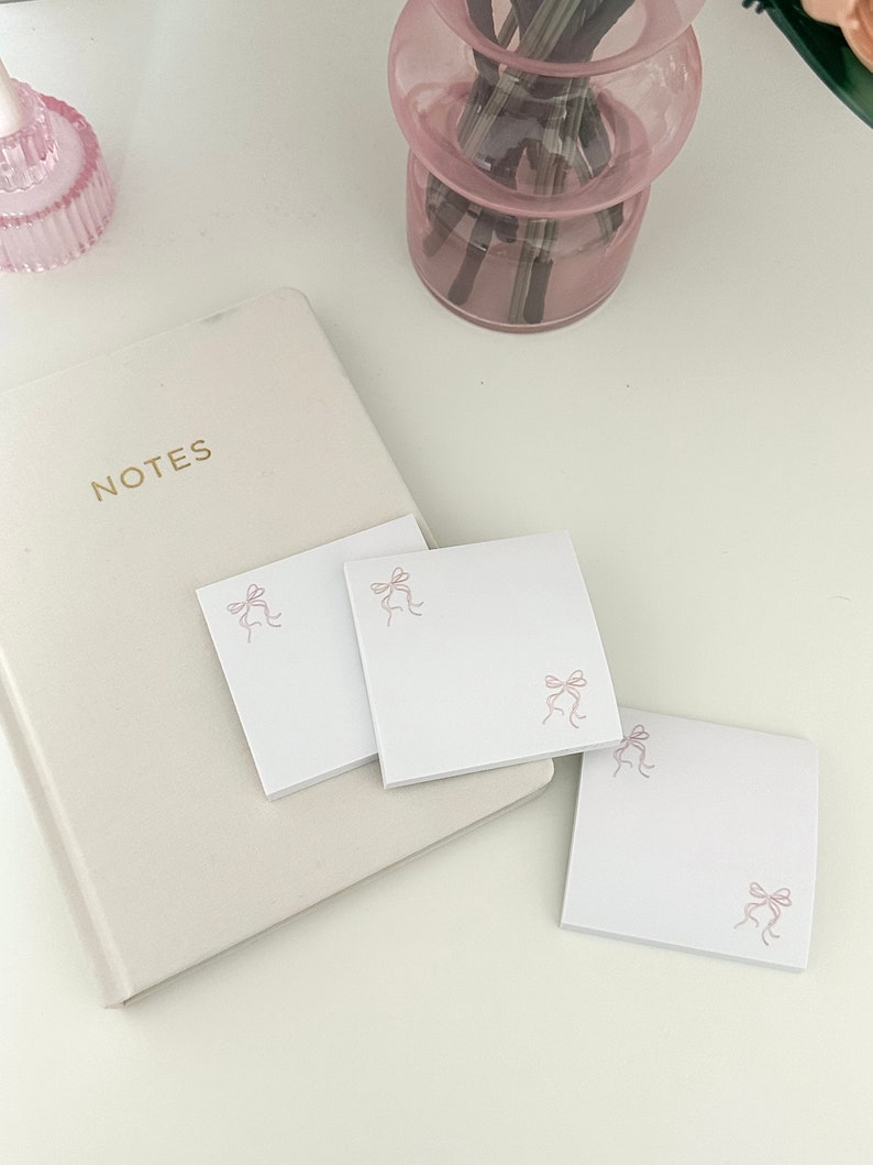 Peut inclure: Trois blocs de notes autocollantes carr&eacute;s blancs avec un motif de n&oelig;ud rose. Les blocs de notes sont sur une surface blanche avec un carnet blanc et un vase en verre rose en arri&egrave;re-plan.