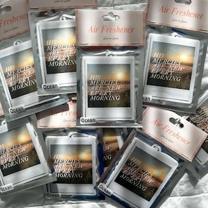 May include: A close-up of several car air fresheners with a sunset scene and the text "His mercies are new every morning". The air fresheners are packaged in clear plastic bags with a white header that says "Air Freshener".