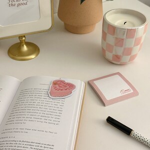 May include: A pink and white checked candle sits on a white surface next to a book with a pink bookmark. The bookmark has a heart shape with embroidery. The book is open to a page with the text "Focus on the good."