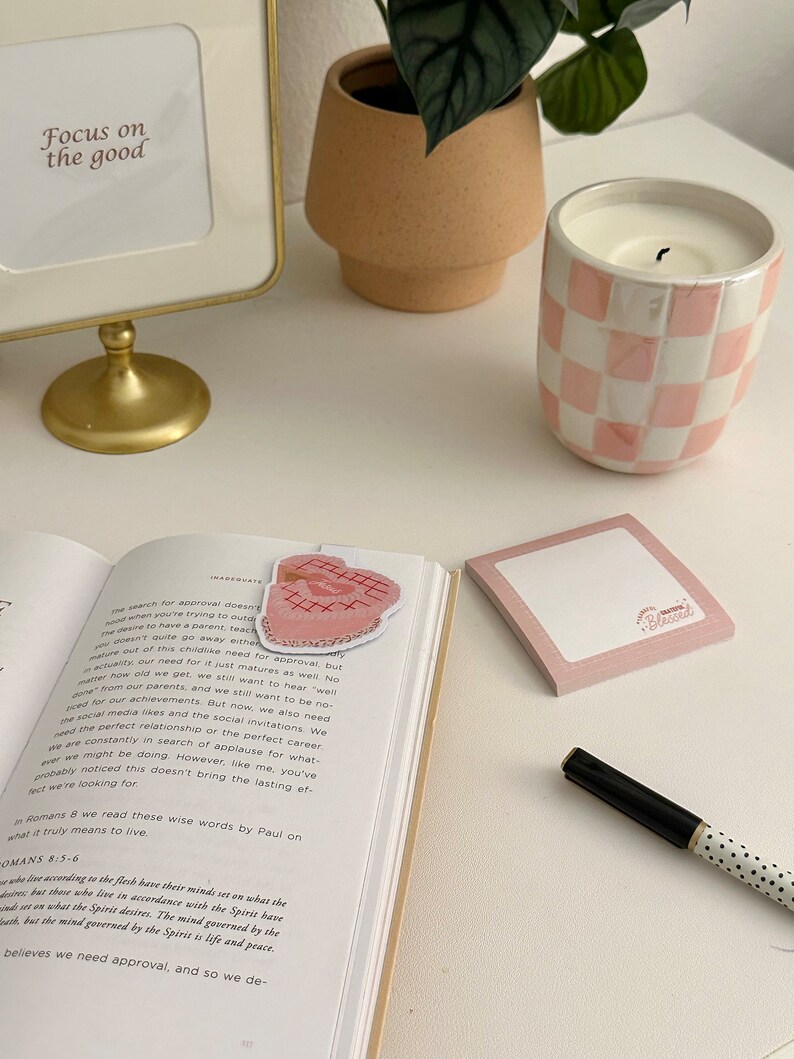 May include: A pink and white checked candle sits on a white surface next to a book with a pink bookmark. The bookmark has a heart shape with embroidery. The book is open to a page with the text "Focus on the good."