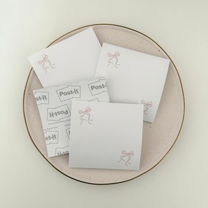 Peut inclure: Quatre notes adh&eacute;sives blanches avec des n&oelig;uds roses sur une assiette rose clair. Les notes adh&eacute;sives portent la marque "Post-it" imprim&eacute;e sur elles.