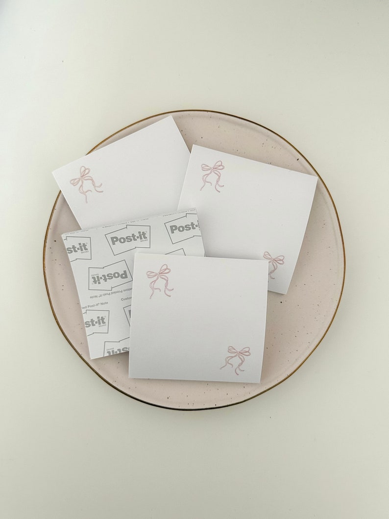 Peut inclure: Quatre notes adh&eacute;sives blanches avec des n&oelig;uds roses sur une assiette rose clair. Les notes adh&eacute;sives portent la marque "Post-it" imprim&eacute;e sur elles.