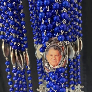 May include: A group of blue rosary beads with silver accents and a silver oval pendant featuring a photo of a man.