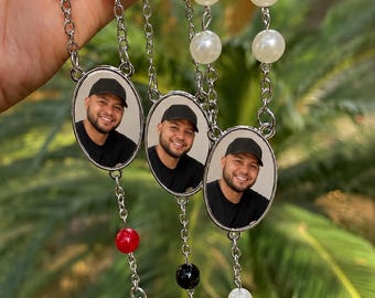 May include: Three rosaries with oval pendants featuring a smiling man in a black cap. The rosaries have red, black, and white beads, with silver chain links and crosses. The background is blurred green foliage.