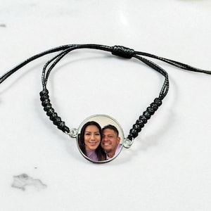 May include: A black adjustable bracelet featuring a round, silver-toned charm with a color photograph of a smiling couple. The bracelet is made of black cord and has a sliding knot closure.