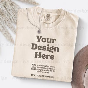 Może przedstawiać: Beżowa koszulka z nadrukiem "Your Design Here" w kolorze brązowym. Pod głównym tekstem znajduje się napis "Add your design with your favorite graphic editing app and add to your store! IVY OLIVER MOCKS".