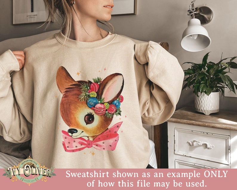 May include: A beige sweatshirt with a vintage-style illustration of a deer wearing a wreath of Christmas ornaments and a pink bow.