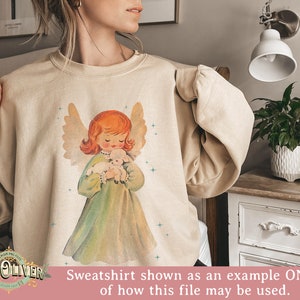 May include: A beige sweatshirt with a vintage-style illustration of an angel holding a lamb. The angel has red hair and is wearing a green dress. The background is a light beige with a subtle sparkle effect.