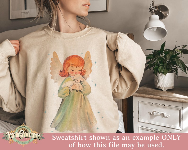 May include: A beige sweatshirt with a vintage-style illustration of an angel holding a lamb. The angel has red hair and is wearing a green dress. The background is a light beige with a subtle sparkle effect.