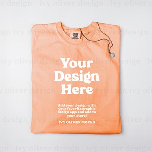 May include: A folded peach-colored t-shirt with the text "Your Design Here" and the words "Add your design with your favorite graphic design app and add to your store! IVY OLIVER MOCKS" printed on the front.