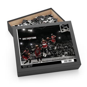 Könnte beinhalten: Ein 500-Teile-Puzzle in einer schwarzen Schachtel mit einer schwarz-weißen Basketballszene mit roten Akzenten. Das Bild zeigt einen Spieler im roten Trikot mit der Nummer 23 in drei Aktionsposen. Text liest GIVE EVERYTHING und 16x20 Puzzle.