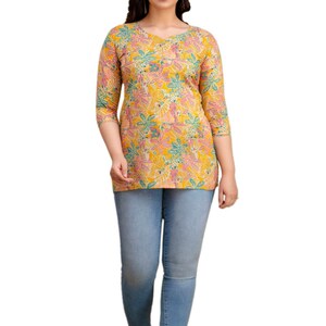 Puede incluir: Una mujer con una blusa amarilla con estampado floral, mangas tres cuartos y cuello en V. Tambi&eacute;n lleva vaqueros pitillo azul claro y sandalias verdes. La blusa tiene un estampado floral colorido.