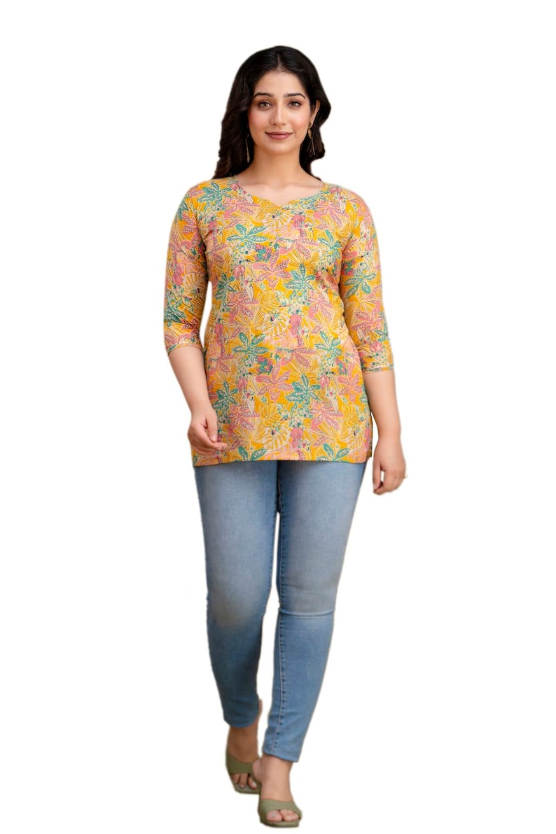 Puede incluir: Una mujer con una blusa amarilla con estampado floral, mangas tres cuartos y cuello en V. Tambi&eacute;n lleva vaqueros pitillo azul claro y sandalias verdes. La blusa tiene un estampado floral colorido.