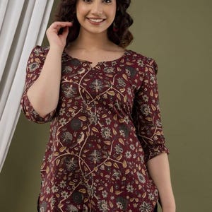 Puede incluir: Top burdeos con estampado floral y mangas tres cuartos. El top presenta un escote redondo y una silueta entallada. El estampado floral incluye detalles en beige y gris. La modelo lleva vaqueros azules.
