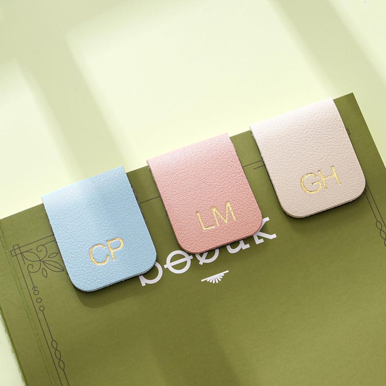 May include: Three pastel-coloured leather magnetic bookmarks, light blue, pink, and cream. Each bookmark features gold lettering with initials. The bookmarks are attached to the pages of a green notebook.