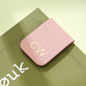 May include: A pink leather bookmark with the embroidered initials "CW" in yellow. The bookmark is placed on a green book with white text. The background is a light yellow-green.