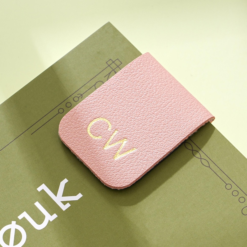 May include: A pink leather bookmark with the embroidered initials "CW" in yellow. The bookmark is placed on a green book with white text. The background is a light yellow-green.