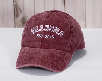 May include: A maroon baseball cap with the word "GRANDMA" embroidered in white, along with "EST. 2015". The cap has a curved brim and a fabric strap closure. The hat is made of a soft, textured material.