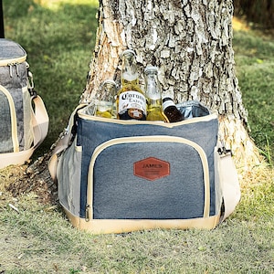 May include: A blue and tan insulated cooler bag with a brown leather patch that says "JAMES" on it. The cooler bag is filled with bottles of beer, including Corona Extra.