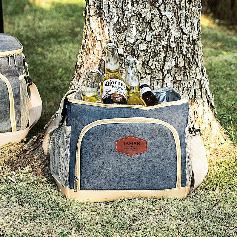 May include: A blue and tan insulated cooler bag with a brown leather patch that says "JAMES" on it. The cooler bag is filled with bottles of beer, including Corona Extra.