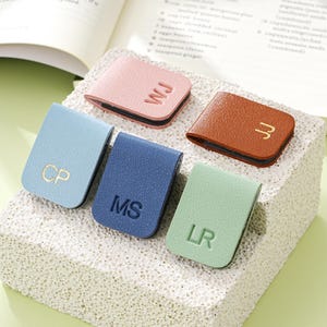 May include: Five leather magnetic bookmarks in shades of pink, brown, blue, light blue, and green. Each bookmark has embossed initials in gold or a matching colour. The bookmarks are displayed on a textured, off-white surface.