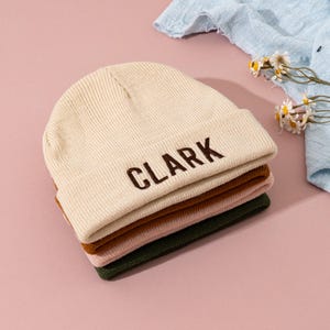 Peut inclure: Une pile de quatre bonnets en tricot de couleurs crème, marron, rose et vert. Le bonnet supérieur, de couleur crème, porte le nom "CLARK" brodé en marron. Les bonnets sont pliés et empilés, prêts à être portés.