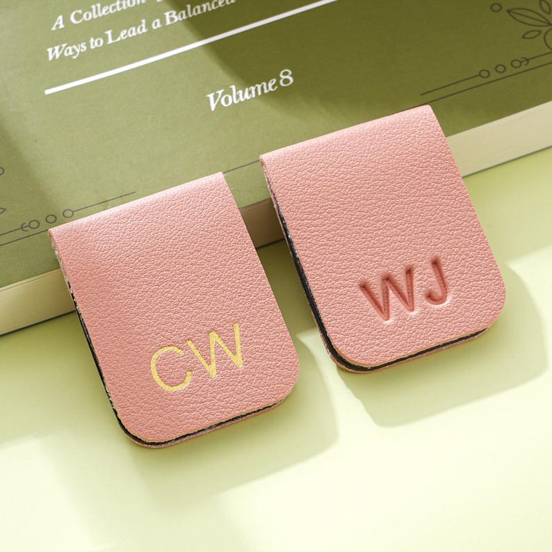 May include: Two pink leather book page holders with gold initials. One has "CW" and the other has "WJ". The book page holders are on top of a green book with the text "A Collection of Ways to Lead a Balanced Life" and "Volume 8".