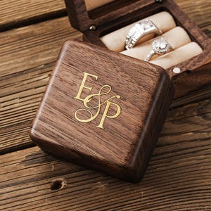 May include: A wooden ring box with gold-coloured initials "ESP" on the lid. The box is open, displaying two rings with silver bands and clear stones. The box is on a wooden surface.