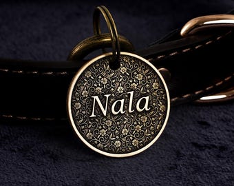 May include: A round, bronze-colored pet tag with the name "Nala" in elegant white script. The tag features a detailed floral pattern and is attached to a black leather collar with bronze hardware. The tag is designed for pet identification.