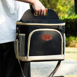 May include: Black and beige insulated cooler bag with a shoulder strap and a top handle. The bag has a mesh pocket on the side and a brown leather patch with the letters "T.S." and crossed golf clubs.
