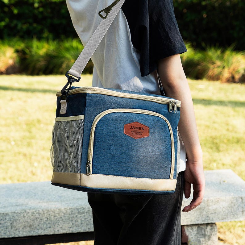 May include: A blue and beige insulated lunch bag with a shoulder strap. The bag has a mesh pocket on the side and a zippered closure. The bag has a brown leather patch with the text "JAMES" on it.