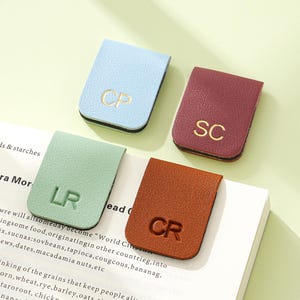 May include: Four leather magnetic bookmarks in pastel shades: light blue, burgundy, mint green, and brown. Each bookmark has gold initials: CP, SC, LR, and CR. The bookmarks are placed on a book.