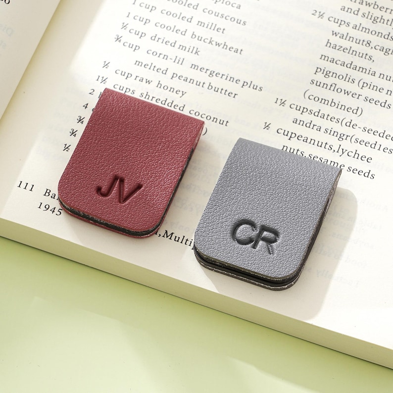 May include: Two leather bookmarks, one claret and one grey, each with initials embossed. The claret bookmark has "JV" and the grey one has "CR". Both are rectangular with rounded corners, resting on a book page.