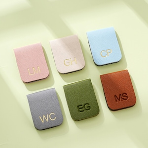 May include: Six leather magnetic bookmarks in shades of pink, beige, light blue, grey, olive green, and brown. Each bookmark has gold initials. The bookmarks are displayed on a light green surface.