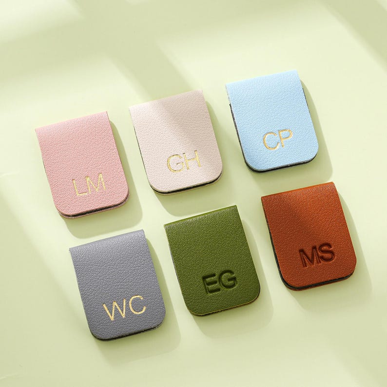 May include: Six leather magnetic bookmarks in shades of pink, beige, light blue, grey, olive green, and brown. Each bookmark has gold initials. The bookmarks are displayed on a light green surface.