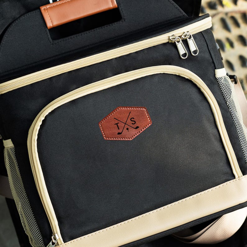 May include: Black and beige insulated cooler bag with a brown leather patch featuring the initials "T.S." and crossed golf clubs.