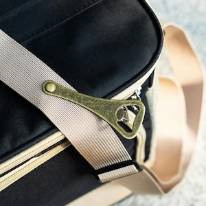 May include: A black canvas bag with a beige strap and a brass bottle opener attached to the strap. The bag has a zipper closure and a beige trim.