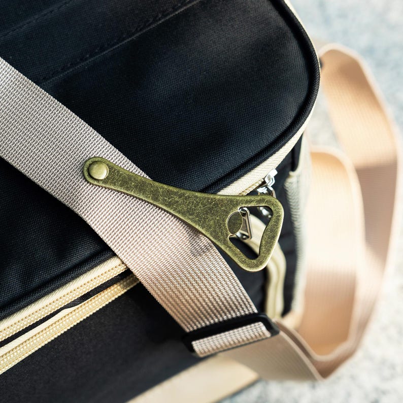May include: A black canvas bag with a beige strap and a brass bottle opener attached to the strap. The bag has a zipper closure and a beige trim.