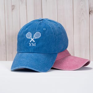 May include: A blue baseball cap with a white embroidered tennis racket design and the initials "YM". A pink visor is partially visible behind the cap. The cap is made of a soft, washed fabric.