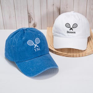 May include: Two baseball caps are shown, one blue and one white. Both caps have a tennis racket design. The blue cap has "YM" embroidered below the rackets. The white cap has "Benson" embroidered below the rackets.