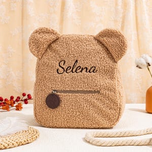 May include: A brown teddy bear-shaped backpack with a zipper closure and a small brown bear charm. The backpack has the name "Selena" embroidered on the front.