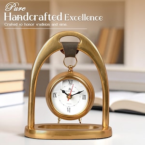 May include: A gold-colored desk clock with a white face and black Roman numerals. The clock is suspended within a gold-colored arched frame and has the text "Pure Handcrafted Excellence" and "Crafted to honor tradition and time."