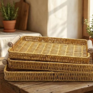 Op de afbeelding: Drie rechthoekige, geweven rieten trays met houten handvatten gestapeld op een rustieke houten tafel. De trays zijn lichtbruin. De achtergrond bevat planten en keukenartikelen.